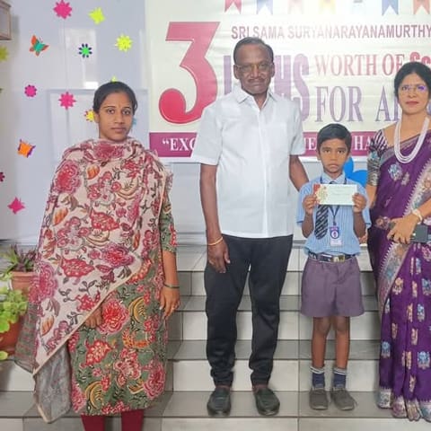 A photograph of four people on a staircase indoors — an older man in a white shirt, a woman in a sari at each side, and a boy in school uniform holding a certificate. Behind them the wall carries a large “35” and the words “In Pursuit of … For All”.